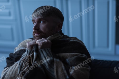 Preview: upset and frozen man looking away while sitting under warm blanket during energy shutdown
