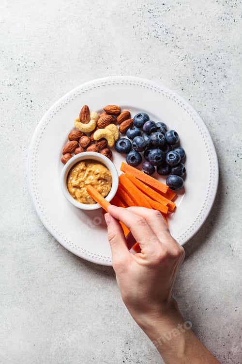 Preview: Woman hand eating healthy snacks plate - carrot, nut, berry and peanut butter, top view.