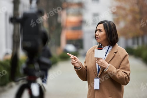Preview: Multiracial female journalist in beige coat reports outdoors during a live news broadcast