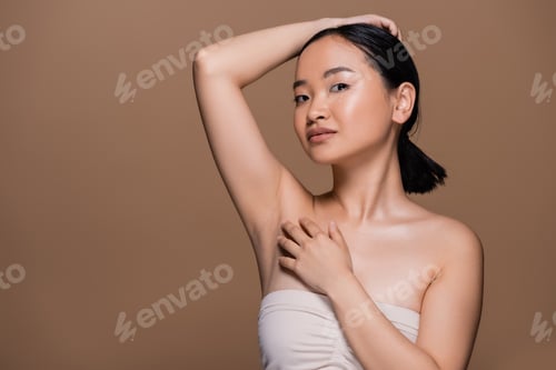 Preview: Pretty asian woman with naked shoulders touching fair and looking at camera isolated on brown