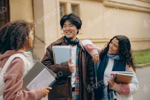 Preview: Young students talking and laughing together outdoor standing near campus and holding books