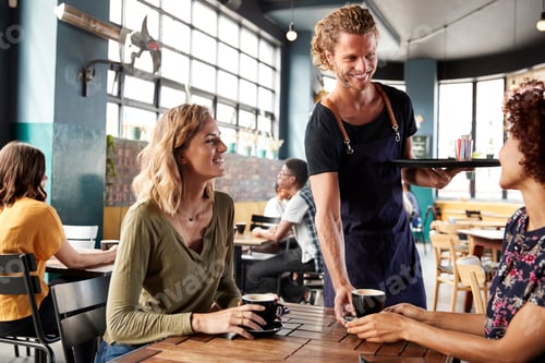 Preview: Two Female Friends Sitting At Table In Coffee Shop Being Served By Waiter