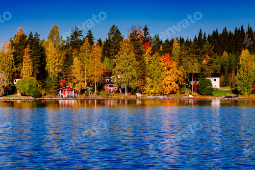 Preview: Autumn foliage, fall colorful forest over blue lake with red cabins in Finland.