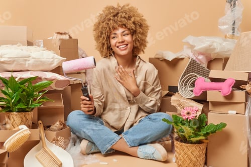 Preview: Happy woman sits crossed legs with painting roller thinks about relocation dressed in shirt and