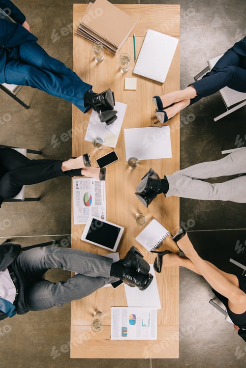 Preview: Top view of legs of resting businesspeople at table with documents and devices in office