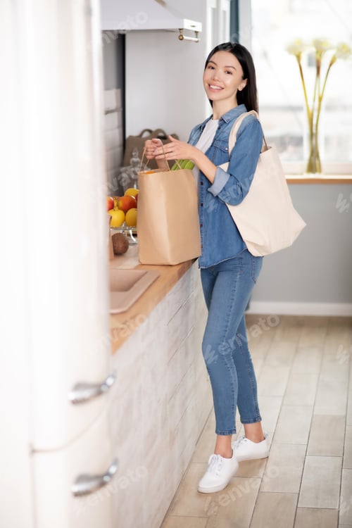 Preview: Young woman standing with shopping bags in the kitchen.