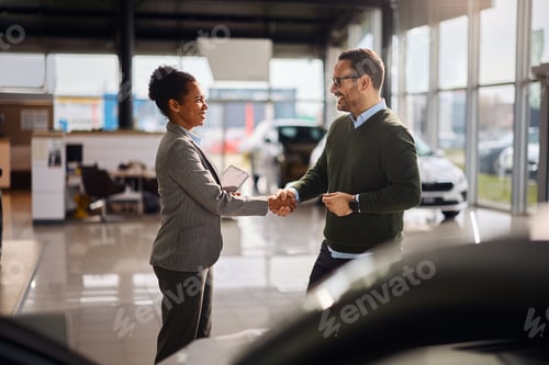 Preview: Happy black saleswoman shaking hands with a customer at car dealership.
