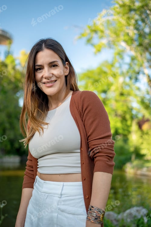 Preview: Smiling woman in white top and brown cardigan standing in a sunny park.
