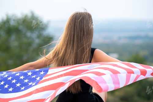 Preview: Young pretty american woman with long hair holding waving on wind USA flag on her sholders standing