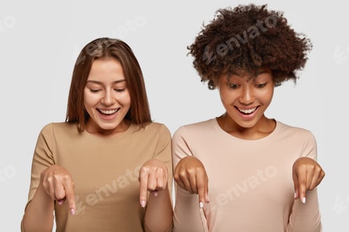 Preview: Horizontal shot of cheerful mixed race two young women point at floor with fore fingers, dressed in