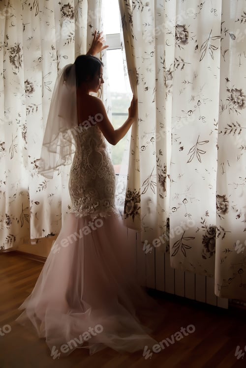 Preview: Cute young woman bride in home white dress at window in country house