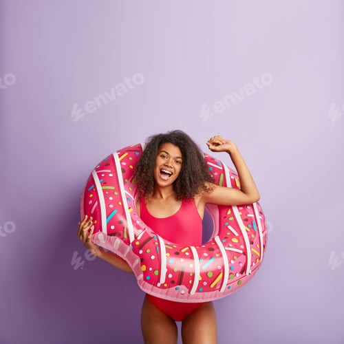 Preview: Funny energetic slim female poses in swimwear inside of donut shaped swim ring, raises hand, rejoice