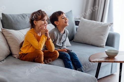 Preview: Two little kids are sitting on grey sofa and watching television.