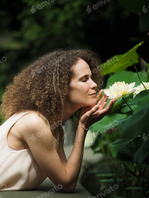 Preview: Young beautiful curly haired woman with closed eyes sniffing lotus flower in botanical garden.