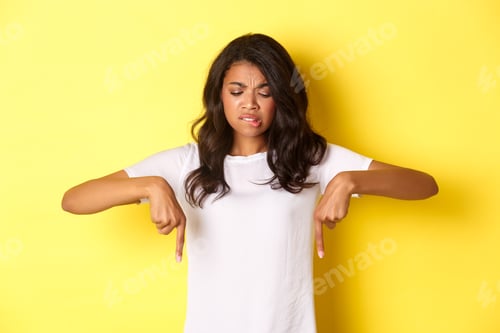 Preview: Portrait of doubtful and skeptical african-american girl in white t-shirt, frowning and pointing