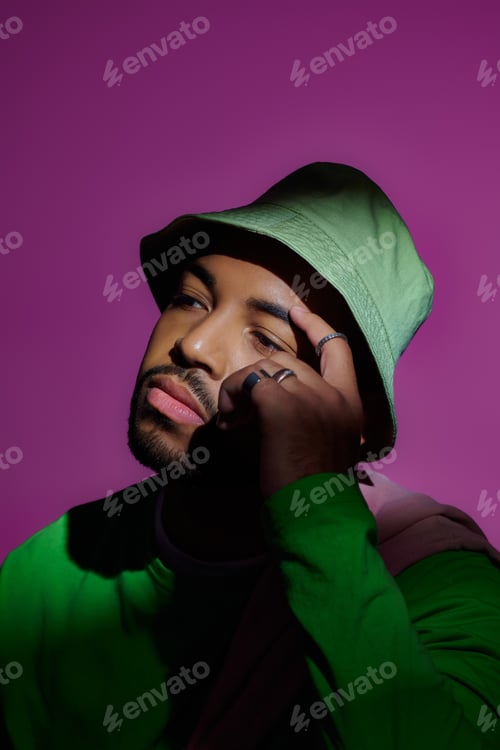 Preview: thoughtful man in green panama with rings touching his brow on purple backdrop, fashion concept
