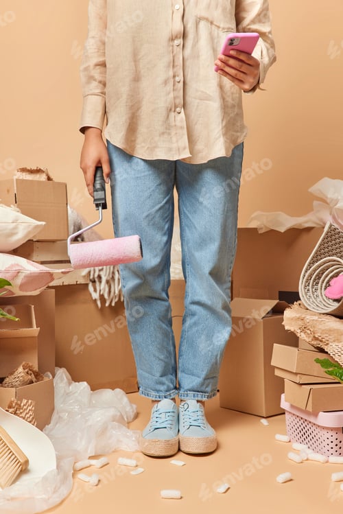 Preview: Cropped shot of faceless woman holds paint roller and smartphone going to make repair of new