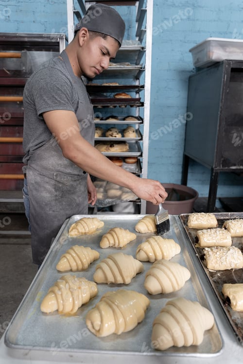 Preview: A young Hispanic baker is brushing egg on some croissants before baking