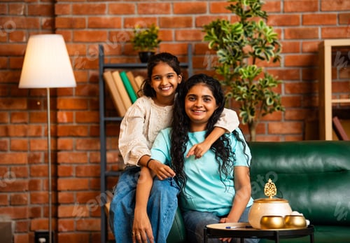 Preview: Playful Indian Asian mother and daughter sitting back to back, having fun on a comfy couch at home