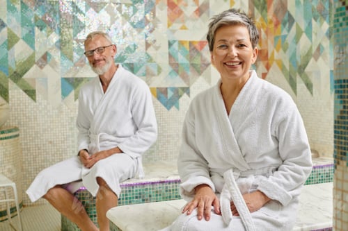 Preview: happy middle aged couple in white robes sitting together in marble sauna, spa wellness concept