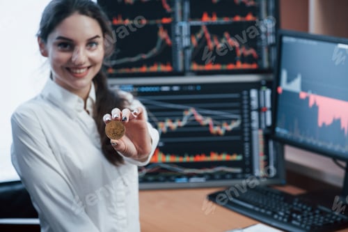 Preview: Beautiful woman holding bitcoin in hands while standing in modern office with many monitors