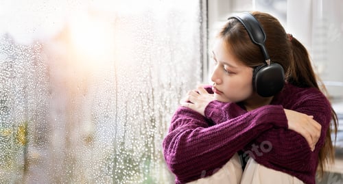 Preview: Depressed young woman with headphone sitting near window at home, closeup