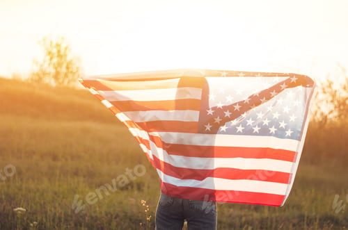 Preview: A young woman holds the US national flag in a field at sunset