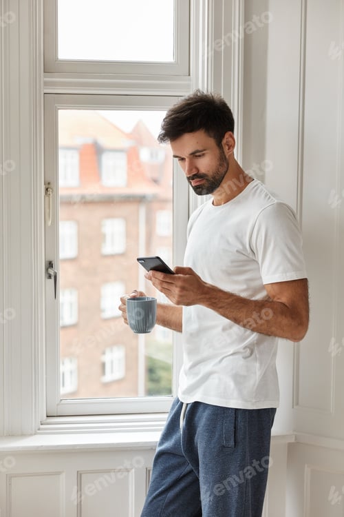 Visualização: Imagem de um belo jovem caucasiano com cerdas grossas, vestido com uma camiseta casual, usa celular novo
