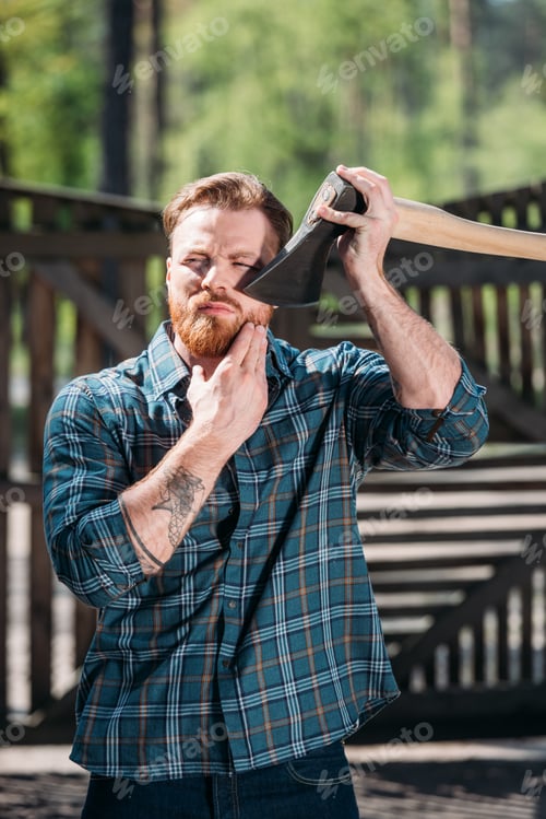 Preview: bearded lumberjack with tattooed hand shaving by axe at sawmill