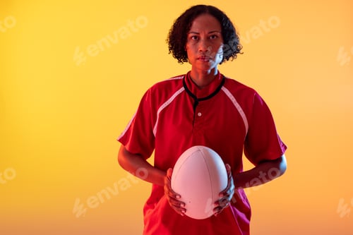 Preview: Portrait of african american female rugby player holding rugby ball with neon yellow lighting