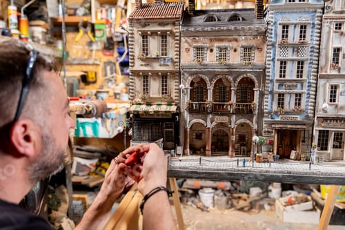 Preview: Modeller man working at scale model of miniature building in his workshop full of tools