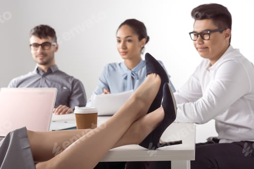 Preview: cropped shot of businesswoman sitting with legs on table while her colleagues working at office
