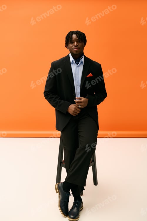 Preview: Handsome young man in stylish black suit poses confidently against bright orange backdrop