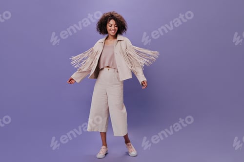 Preview: Charming woman in beige costume dancing on purple background. Happy curly girl in pants and jacket