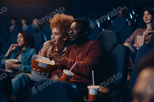 Preview: Happy multiracial couple eating popcorn during movie projection at cinema.