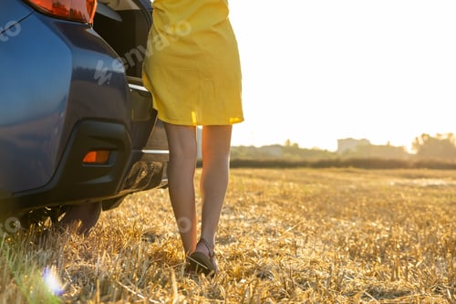 Preview: Back view of young woman driver slim legs in yellow summer dress standing near her car