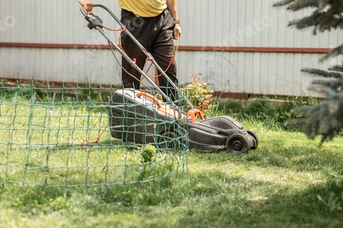 Preview: faceless man mows the grass with an electric lawn mower in a private garden on a clear sunny day