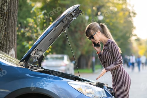 Preview: Young upset woman driver talking on mobile phone near a broken car with open hood