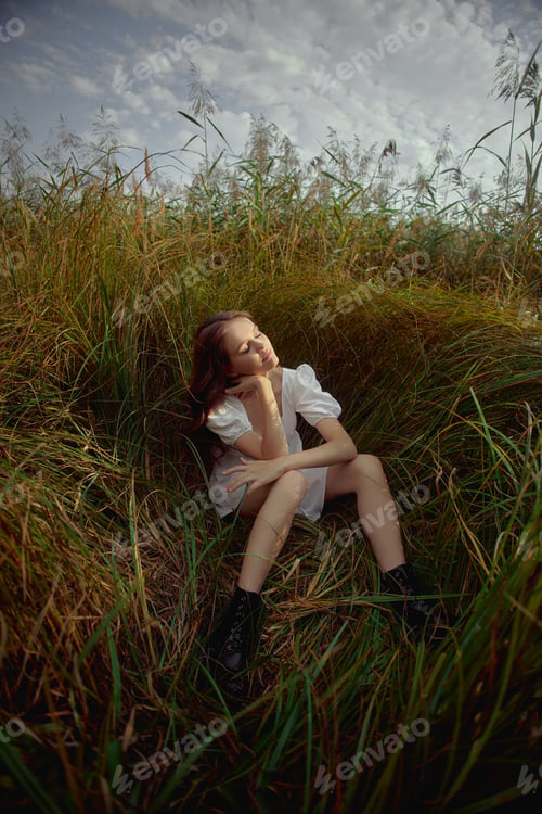 Preview: Beautiful young woman in white summer dress is sitting in tall grass in rural field. Portrait