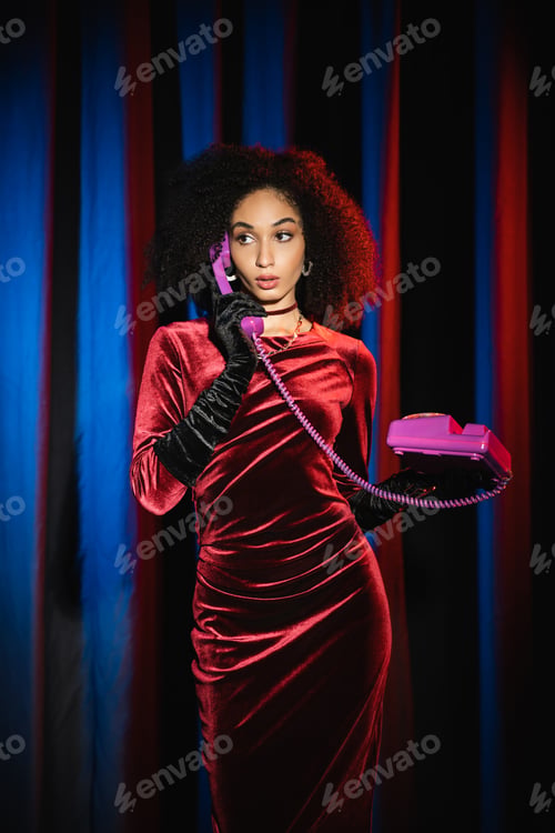 Preview: Stylish african american model in velvet dress talking on telephone near background with blue light
