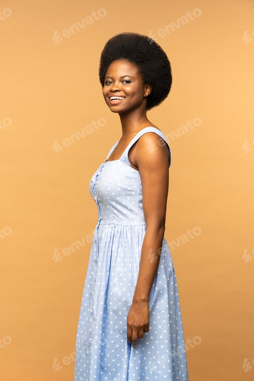 Preview: Pretty Afro American female with afro hairstyle in dress smiling isolated on beige studio background