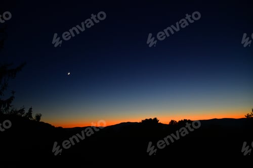Preview: Sliver of moon with few stars in night sky right with glow of the sunset on the horizon of mountains