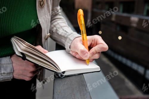 Preview: Woman writing notes in notebook with orange pen outdoors