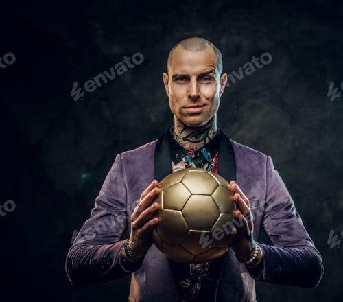 Preview: Elegant tattooed male model posing with a golden soccer ball in a studio
