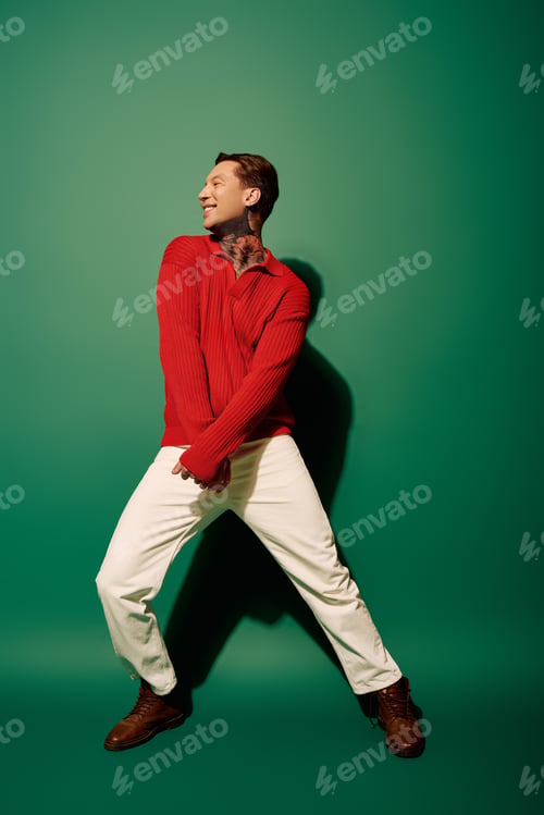 Preview: Young man with tattoos joyfully poses against a vibrant green backdrop in stylish attire