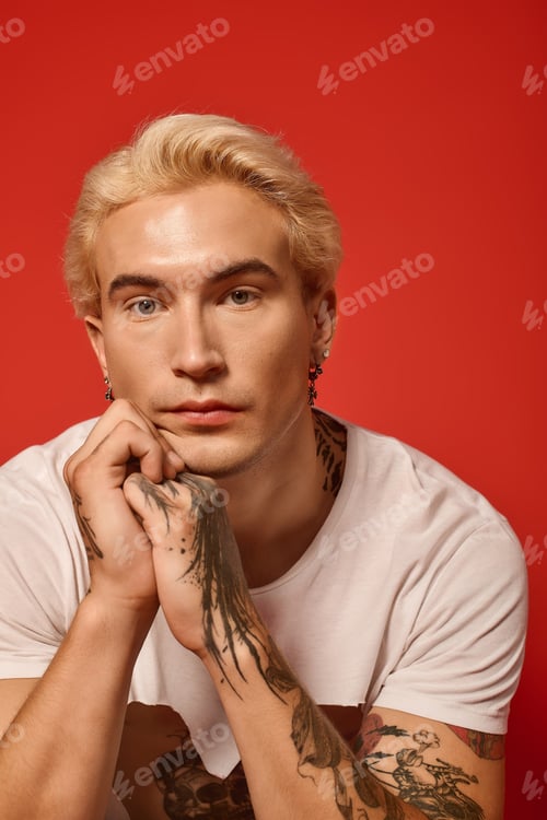 Preview: Stylish young man with blond hair and tattoos poses against a vibrant red backdrop