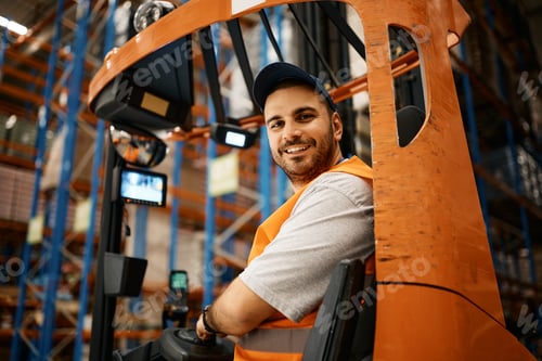 Preview: Happy male worker driving forklift at distribution warehouse and looking at camera.
