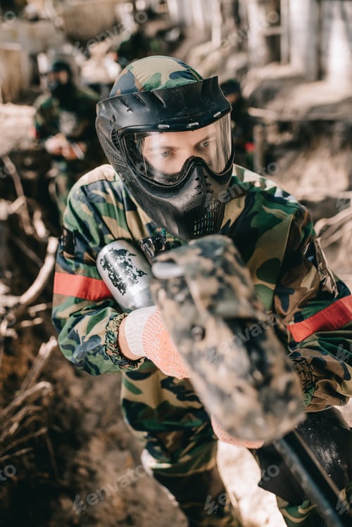 Preview: selective focus of male paintball player in goggle mask and camouflage aiming by paintball gun from