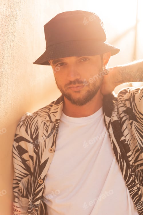 Preview: Hipster man with fashion shirt and hat stands near wall ta sunset. Summer male clothes look