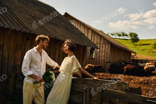 Preview: happy asian bride in wedding dress standing near groom in sunglasses near stable with sheep